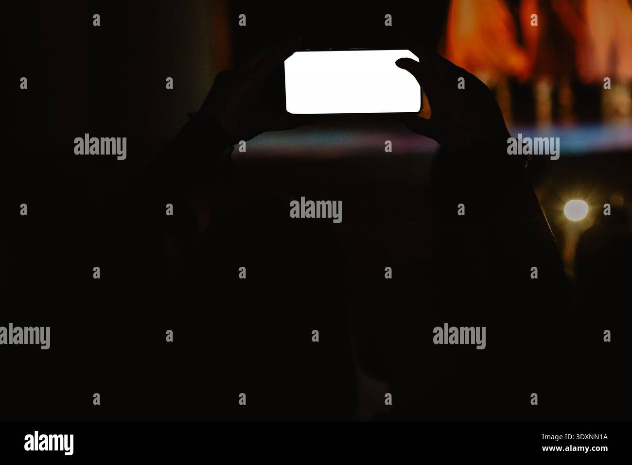 Silhouette d'une personne tenant un smartphone pour enregistrer un concert live ou une performance sur scène la nuit. L'écran lumineux contraste avec la foule sombre, capturant Banque D'Images