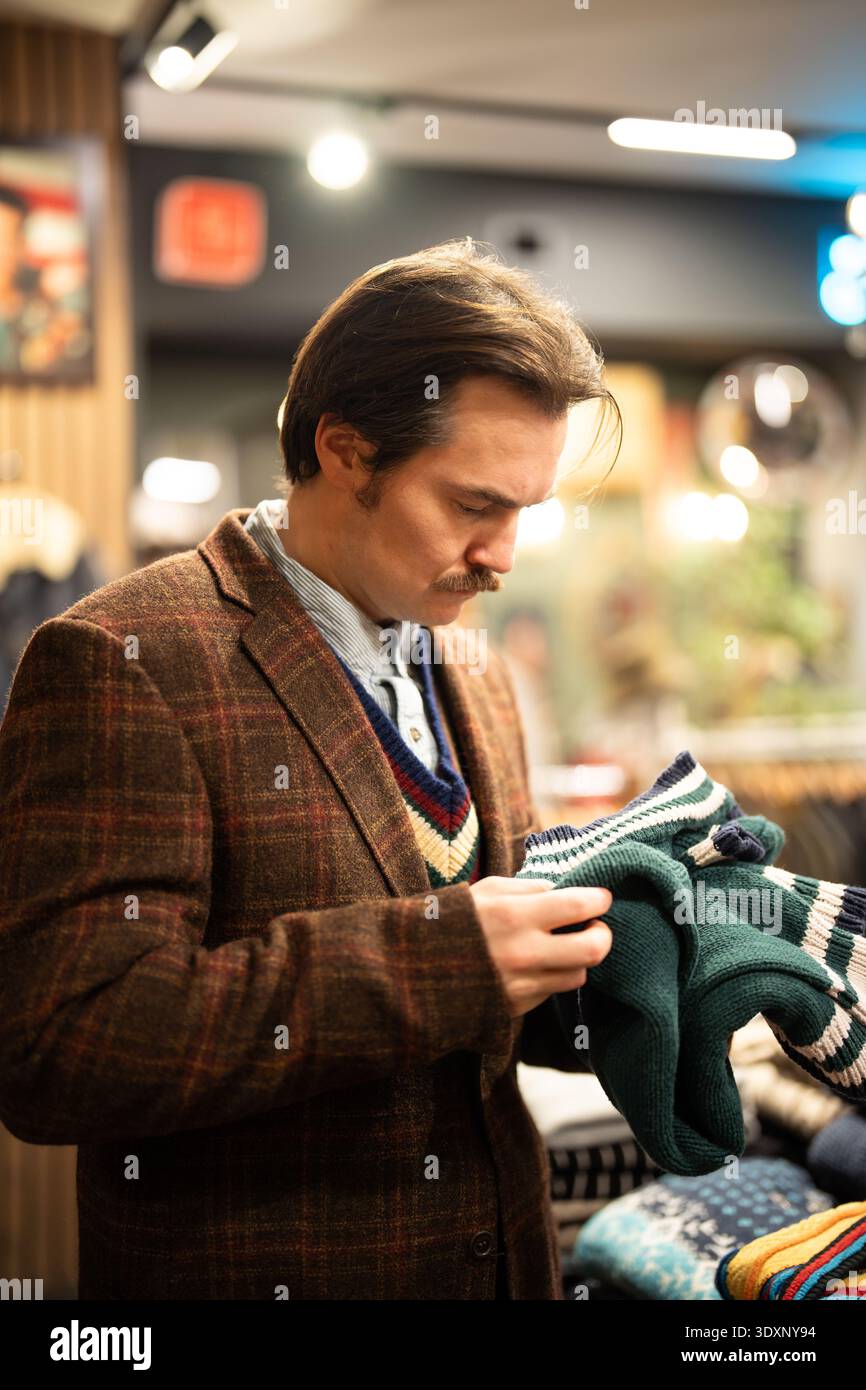 Homme à la mode achetant des vêtements dans la boutique vintage d'occasion, vérifiant les tricots à l'ancienne. Banque D'Images
