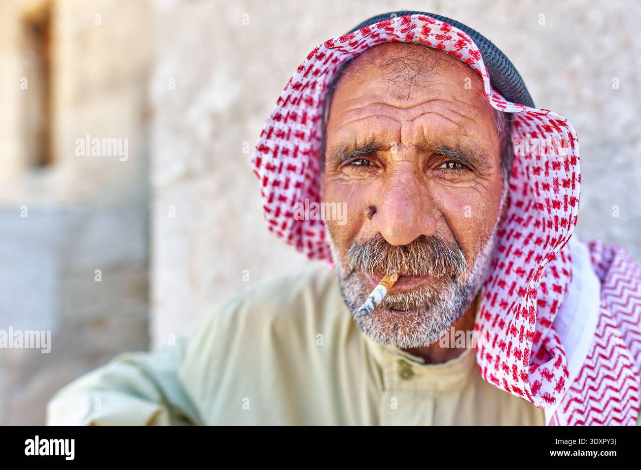 Un homme âgé est assis à Alep, en Syrie, portant des vêtements traditionnels et une écharpe rouge et blanche pendant qu'il fume une cigarette. Banque D'Images