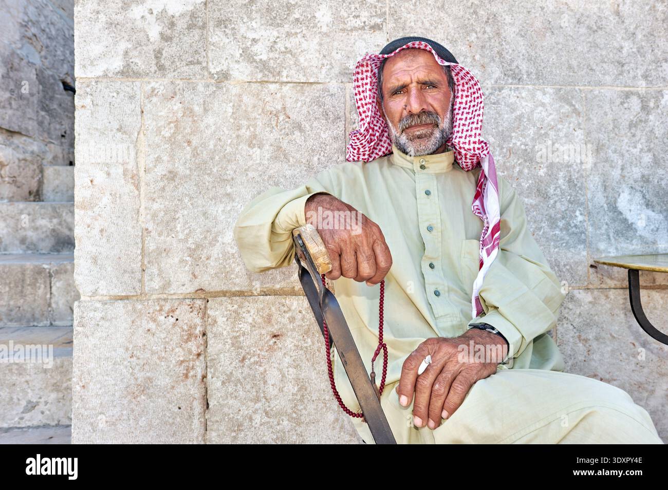 Un homme âgé est assis contre un mur de pierre à Alep. Il porte des vêtements traditionnels et tient un bâton de marche. Banque D'Images