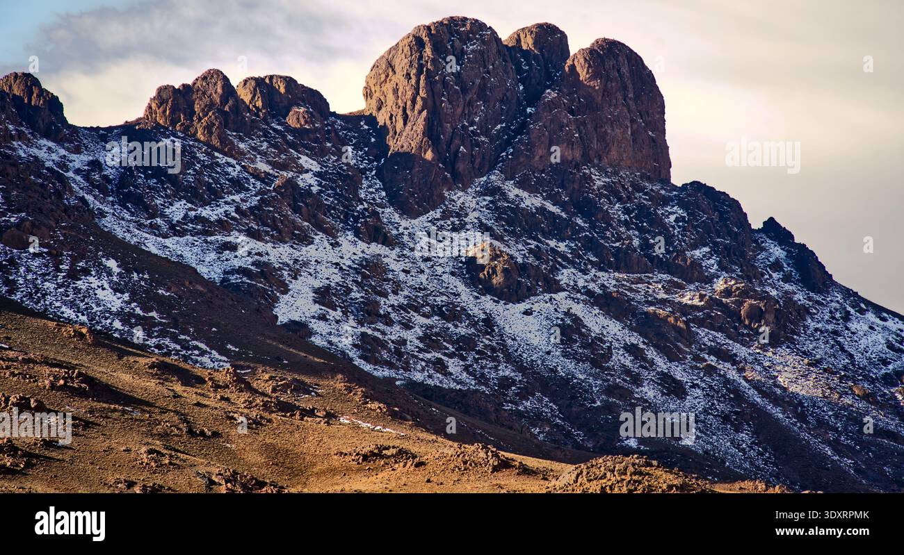 Montagnes de l'Atlas formation rocheuse escarpée s'élevant contre le ciel, montrant des pentes abruptes partiellement couvertes de taches de neige sous la chaude soirée ou le matin Banque D'Images