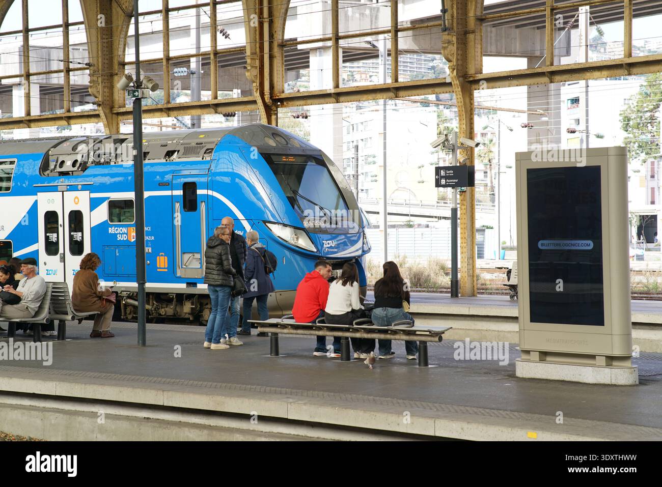 Le train régional Blue Zou attend sur le quai historique de la Gare de Nice ville pendant que les passagers s'assoient sur des bancs et discutent sous la verrière en fer ornée.Nice Banque D'Images