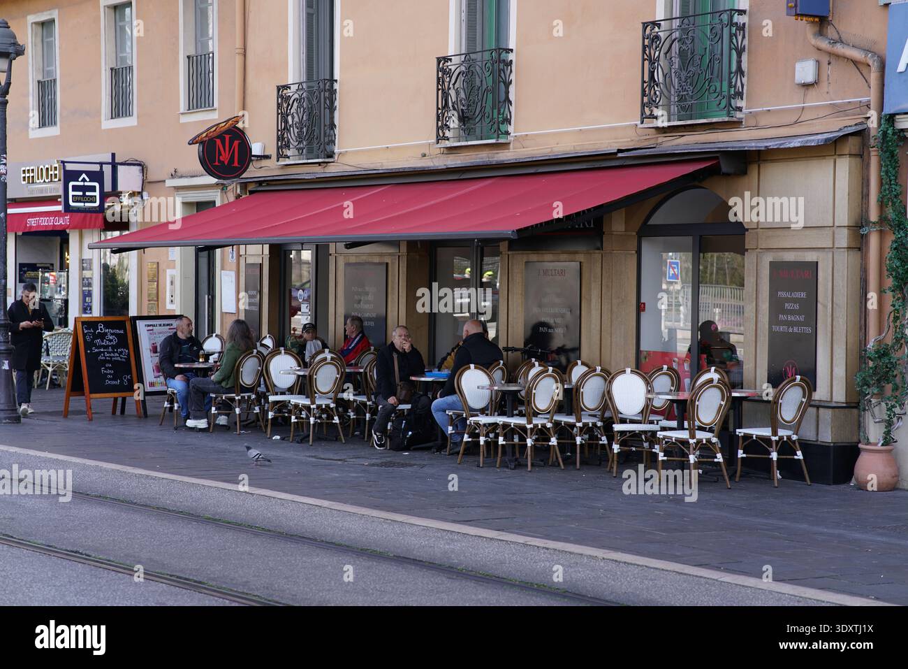 Terrasse extérieure d'un café à Nice avec auvent rouge, chaises en osier, les gens assis à des tables le long de la rue trackée de tram.Nice, France Banque D'Images