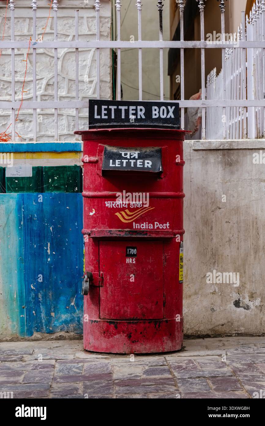 Boîte de pilier Red India Post debout sur une rue pavée à Leh devant une clôture en métal blanc et un mur avec de la peinture bleue et verte. Banque D'Images