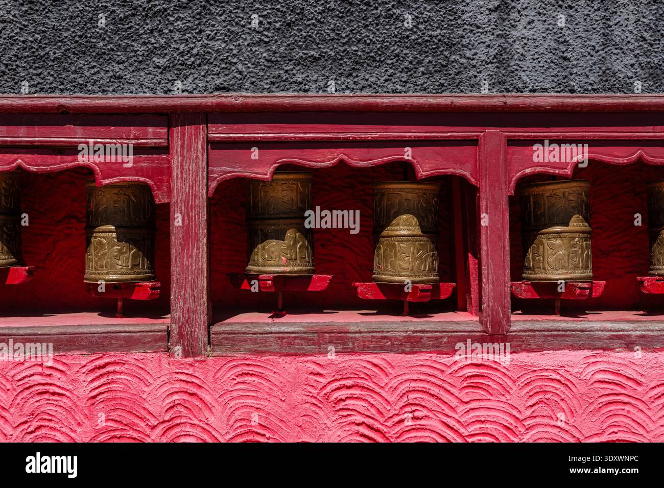 Gros plan de roues à prières dorées avec des inscriptions tibétaines placées dans un cadre en bois rouge contre un mur gris texturé à Thiksey Gompa. Banque D'Images