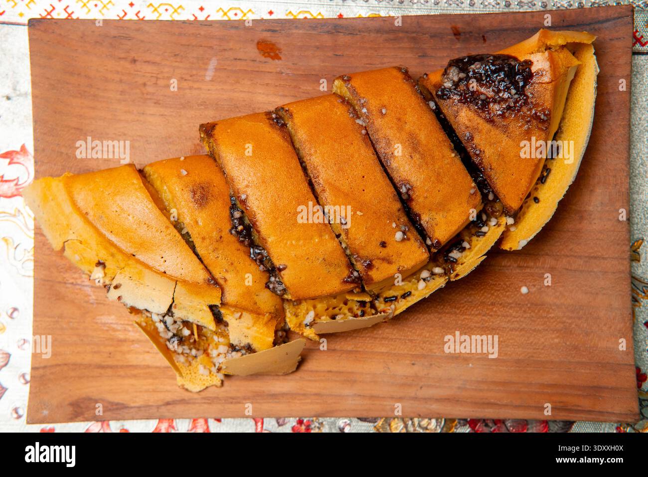 Vue latérale rapprochée de Martabak Manis tranché sur une assiette en bois, montrant une texture épaisse et une garniture de chocolat fondu sur un fond à motifs. Banque D'Images