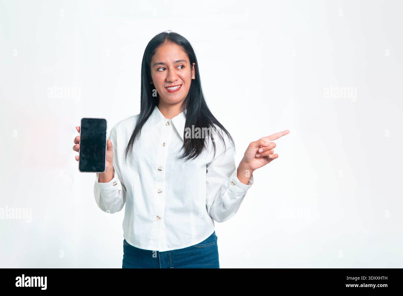 Portrait d'une femme indonésienne souriante dans une chemise blanche professionnelle tenant un téléphone portable avec un écran noir vierge et pointant son doigt vers le s. Banque D'Images