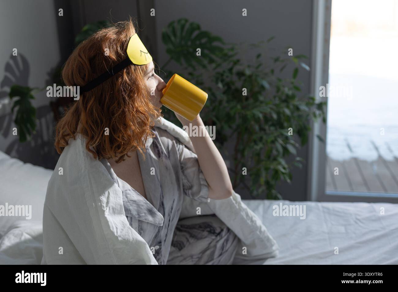 Une femme est confortablement assise sur un lit tout en portant un masque de sommeil et en profitant d'une tasse de café apaisante de sa tasse préférée Banque D'Images