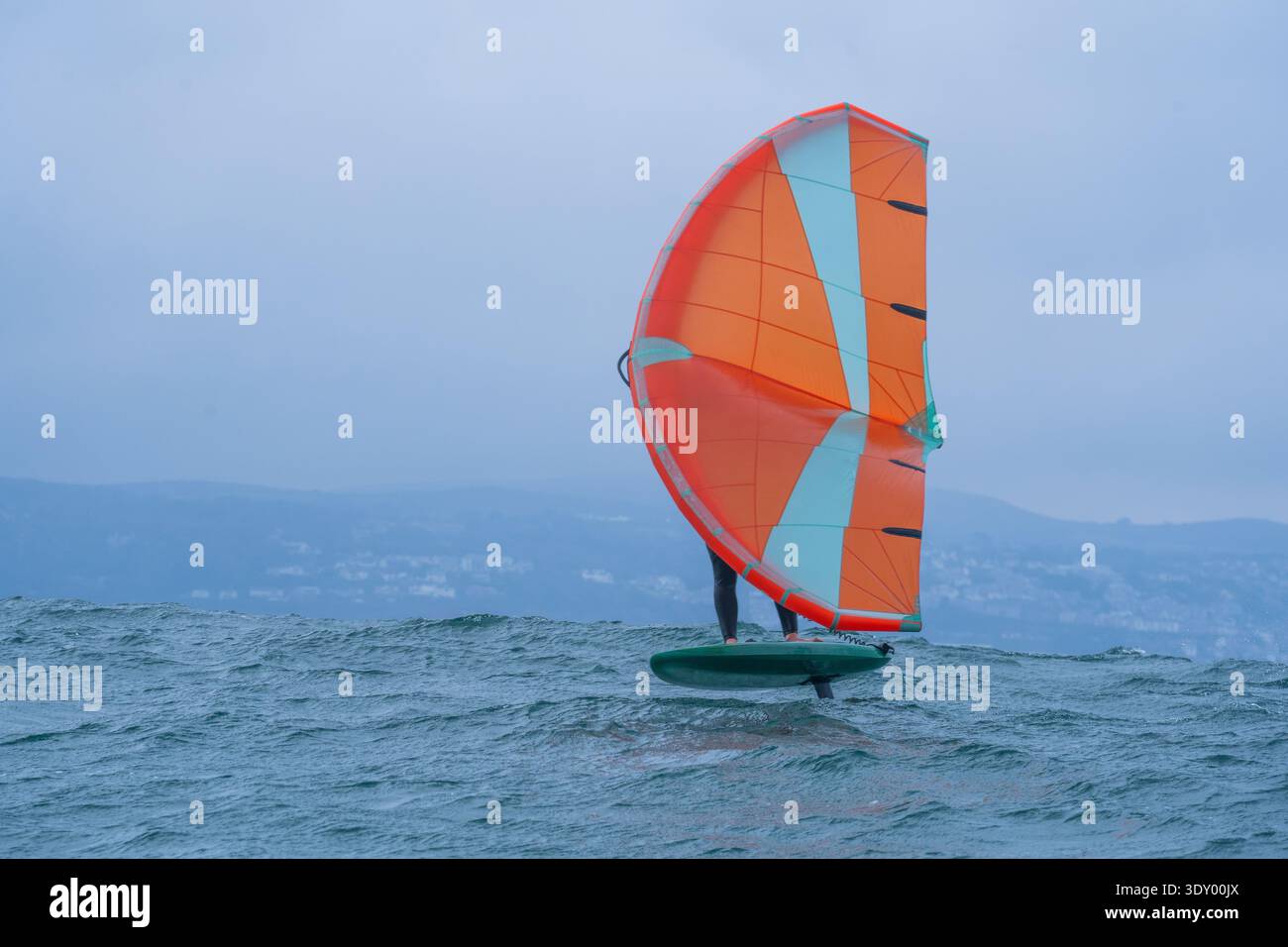 Une aile homme défile en mer à l'aide d'une aile gonflable tenue à la main orange vif, chevauchant une planche de surf hydrofoil. Mer verte, ciel bleu nuageux. Littoral. Banque D'Images