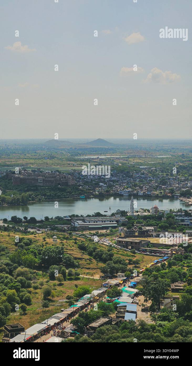 Vue panoramique d'une ville indienne historique entourée de verdure et d'un lac calme, vue du sommet d'une colline sous un ciel bleu clair. Temples, forts et denses. - Image de stock capturée avec un smartphone
