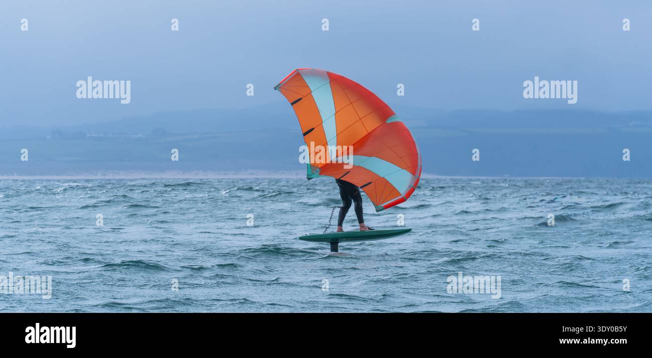 Une aile homme défile en mer à l'aide d'une aile gonflable tenue à la main orange vif, chevauchant une planche de surf hydrofoil. Mer verte, ciel bleu nuageux. Littoral. Banque D'Images