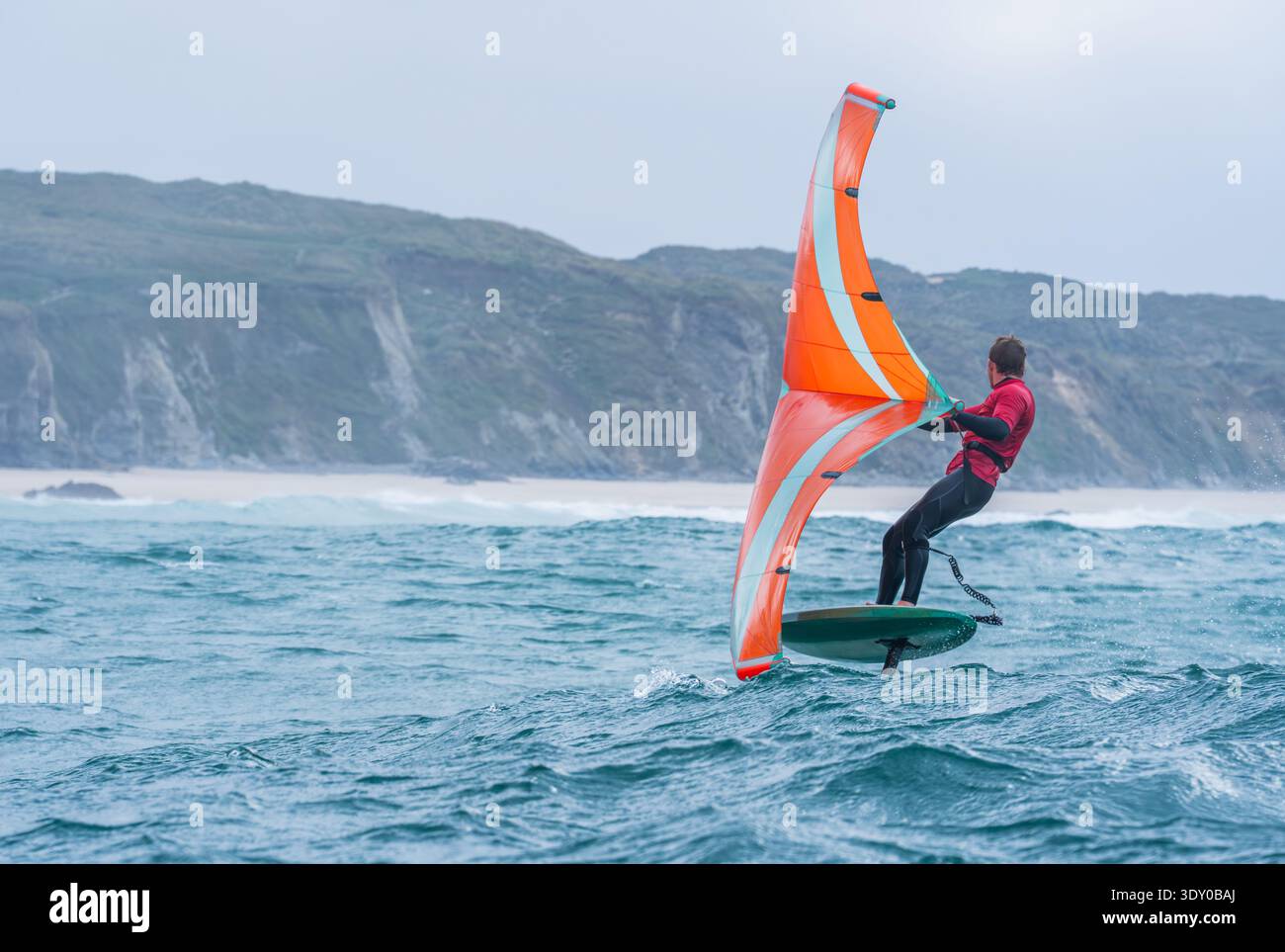 Une aile homme défile en mer à l'aide d'une aile gonflable tenue à la main orange vif, chevauchant une planche de surf hydrofoil. Mer verte, ciel bleu nuageux. Falaises côtières. Banque D'Images