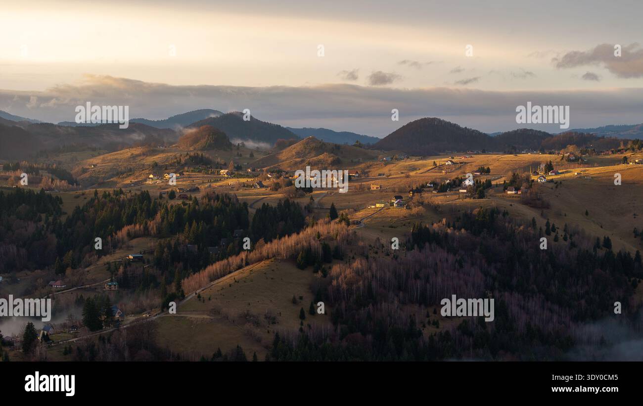 Vue aérienne de la lumière dorée du soleil embrassant les collines et les maisons dispersées, peignant un paysage serein dans des teintes chaudes, Șirnea, Brașov, Roumanie. Banque D'Images