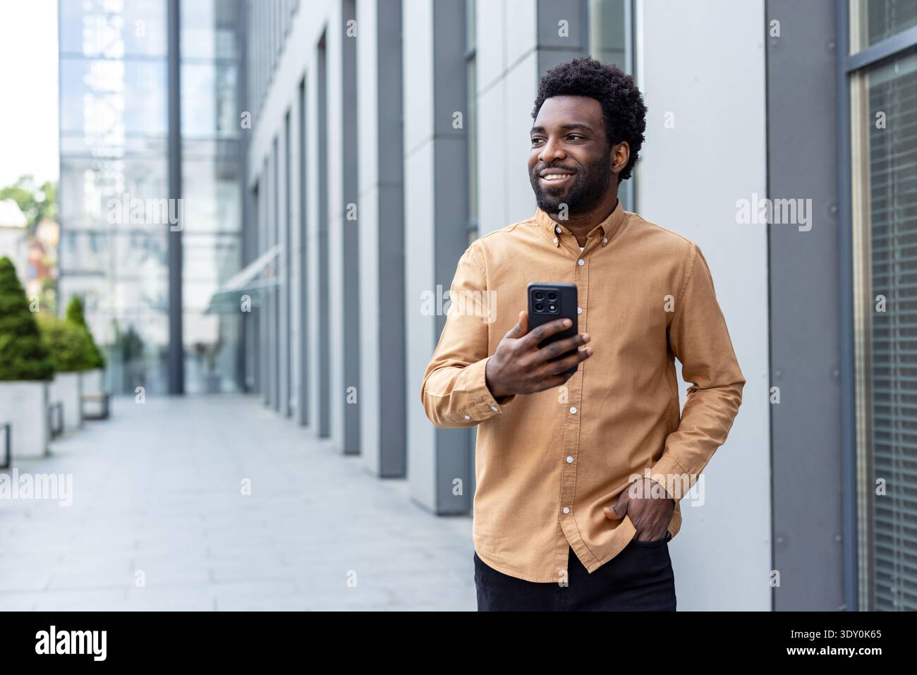 Jeune homme debout à l'extérieur à côté d'un immeuble de bureaux moderne, tenant un téléphone portable et regardant loin avec une expression heureuse et confiante, représentant le succès et le style de vie urbain Banque D'Images