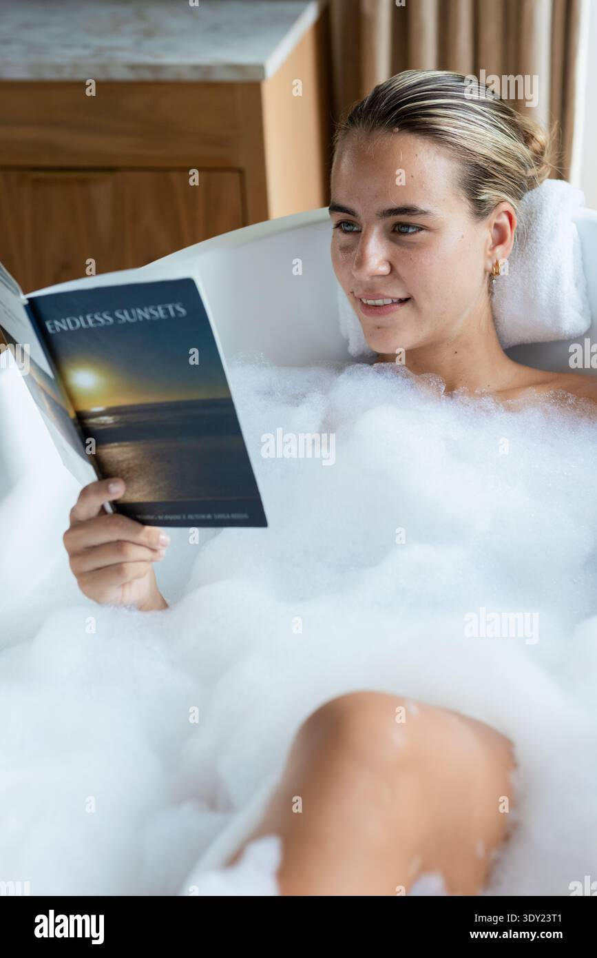 Femme couchée dans une baignoire blanche à la maison avec de la mousse, la tête sur la serviette tenant des couchers de soleil sans fin de livre de poche. Baignoire, bulles, lecture, autosoin, serein, ambiance Banque D'Images