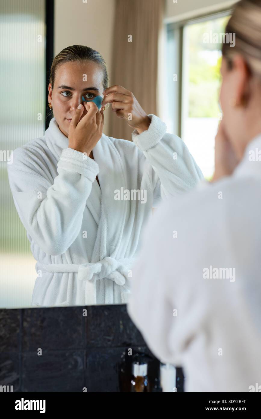 Femme portant un peignoir blanc à la vanité de la salle de bains, appliquant un patch de gel bleu sous les yeux dans le miroir. Femme, soins de la peau, autosoin, réflexion, lumière du jour, vent Banque D'Images