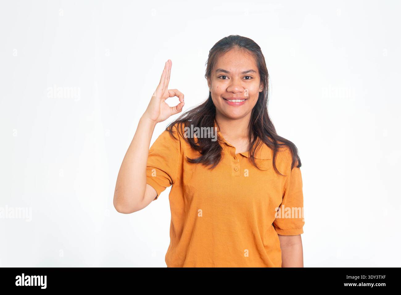 Heureuse femme indonésienne en chemise orange montrant un signe correct avec sa main, souriant à la caméra sur un fond blanc propre. Banque D'Images