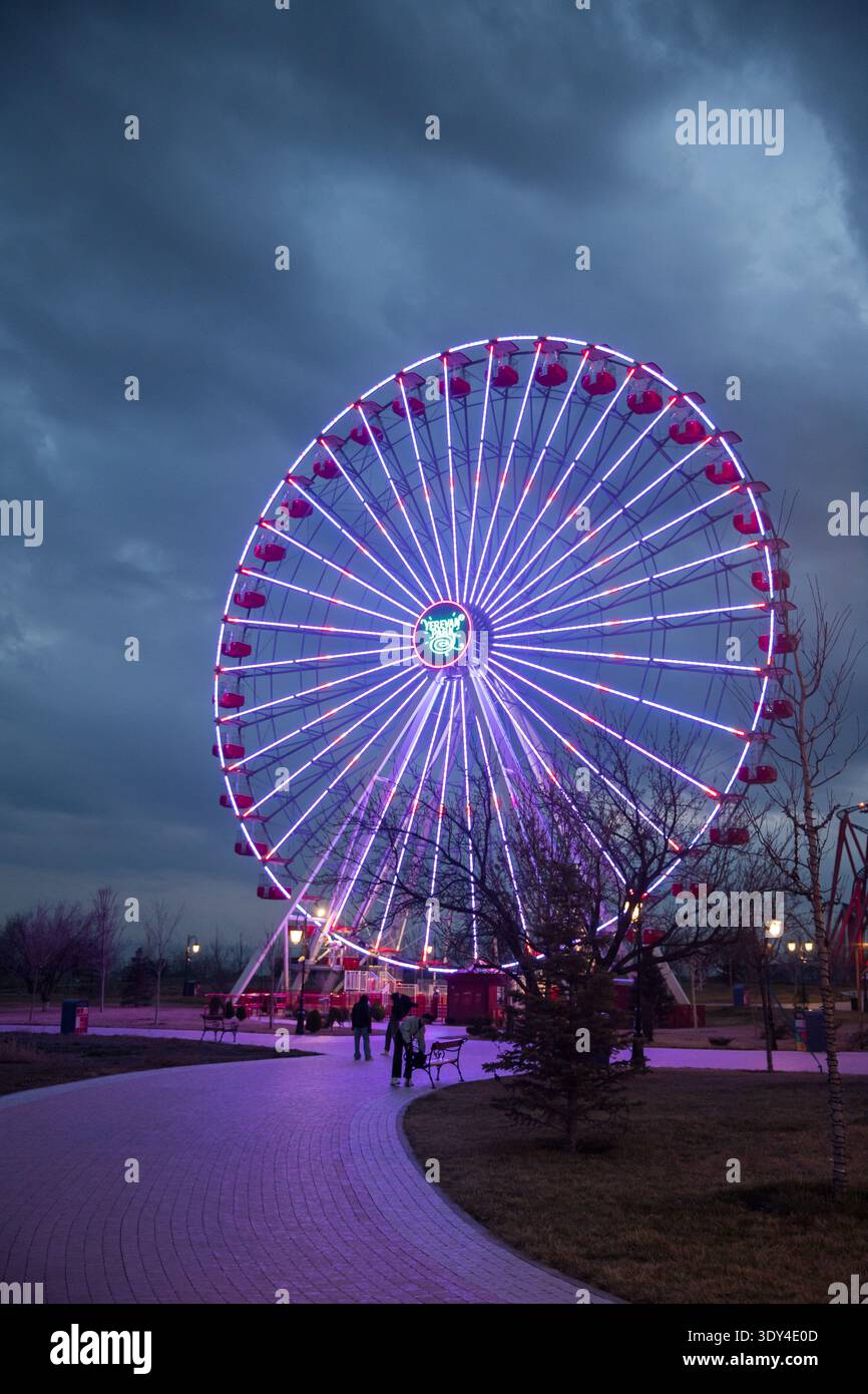 Grande grande roue éclairée au parc d'attractions Erevan Park en soirée. Les gens marchant dans la zone du parc. Banque D'Images
