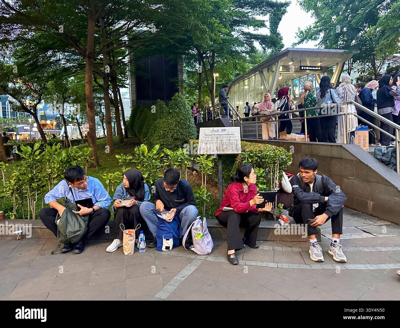 Jakarta - 5 mars 2026 : les amis et les navetteurs s'assoient près d'une entrée de métro partageant de la nourriture et se relaxant dans un petit parc pendant le trajet en soirée. - Image de stock capturée avec un smartphone
