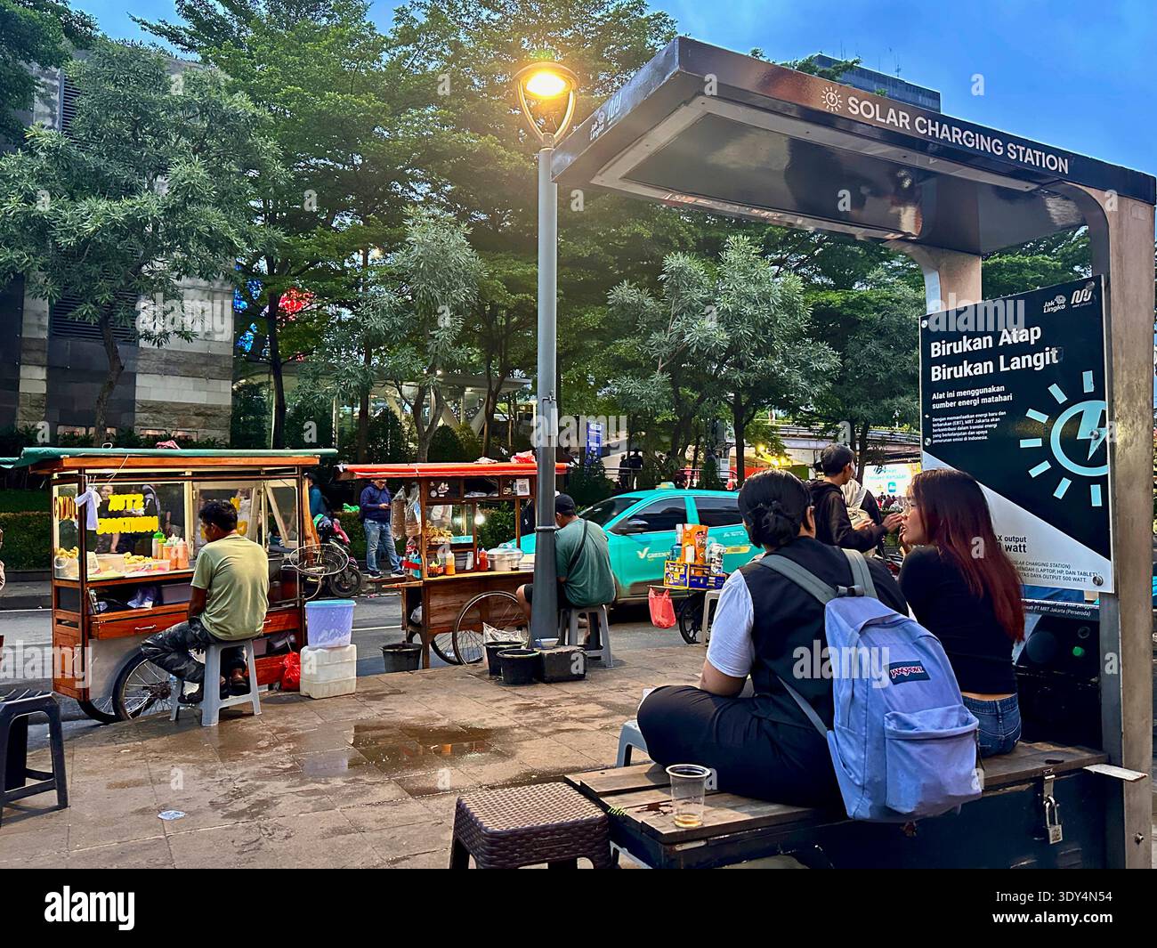 Jakarta - 5 mars 2026 : les gens se rassemblent sur des chariots de nourriture en bord de route à côté d'une station de recharge solaire dans une scène de rue animée en soirée dans le centre de Jakarta. - Image de stock capturée avec un smartphone