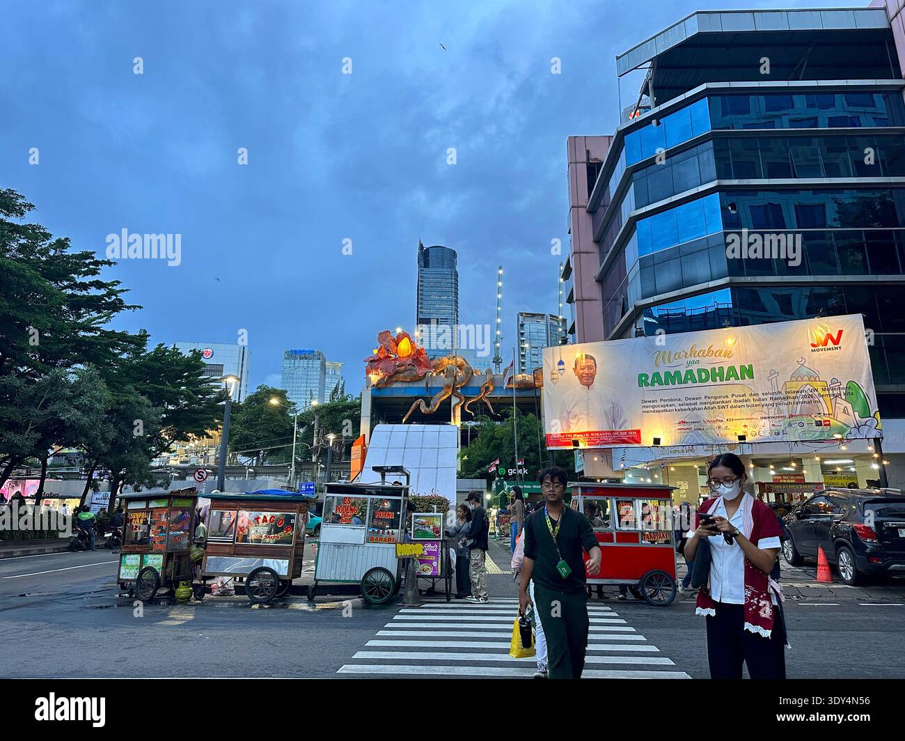 Jakarta - 5 mars 2026 : les piétons traversent une passerelle zébrée près des chariots de nourriture et un panneau d'affichage du Ramadan sous les gratte-ciel du centre de Jakarta. - Image de stock capturée avec un smartphone
