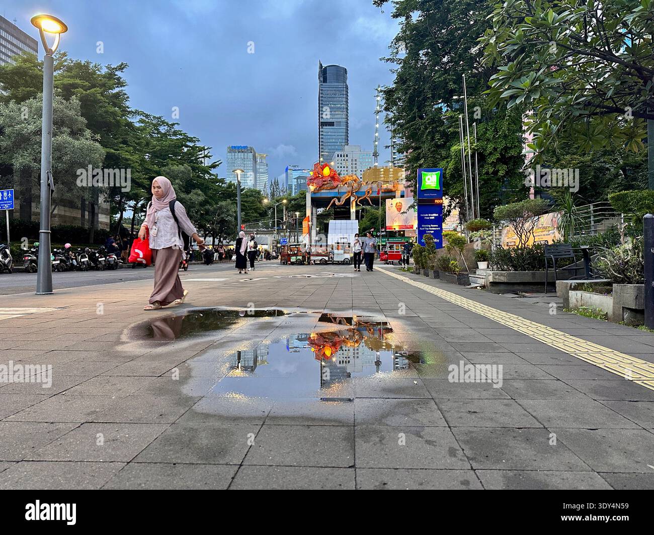 Jakarta - 5 mars 2026 : Une femme dans un hijab marche le long d'une rue animée au crépuscule alors que les flaques reflètent les lumières de la ville et les gratte-ciel dans le centre de Jakarta. - Image de stock capturée avec un smartphone