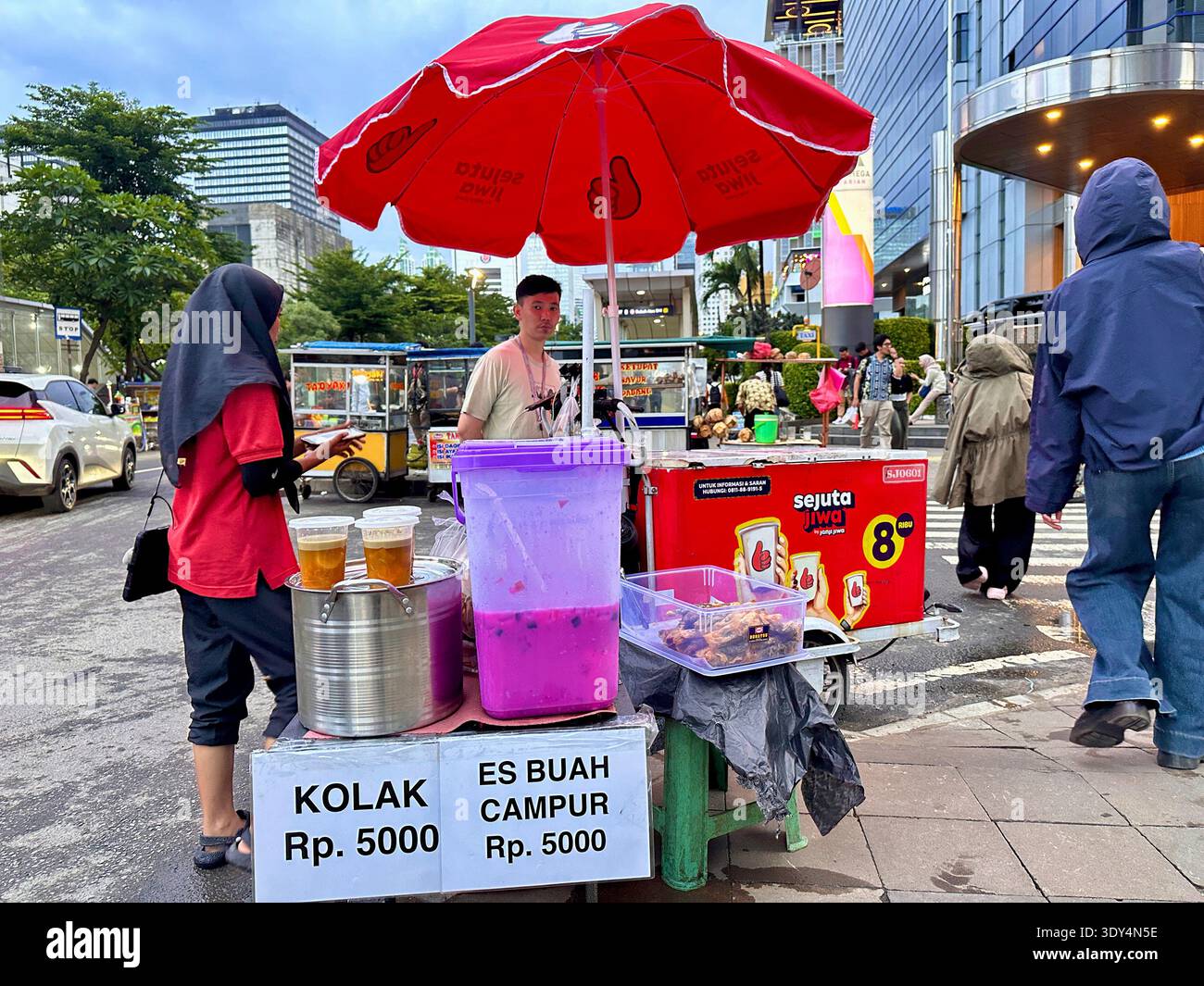 Jakarta - 5 mars 2026 : Un vendeur de rue vend kolak et es buah dans un chariot sous un parapluie rouge sur un trottoir animé près de Bundaran HI au crépuscule. - Image de stock capturée avec un smartphone