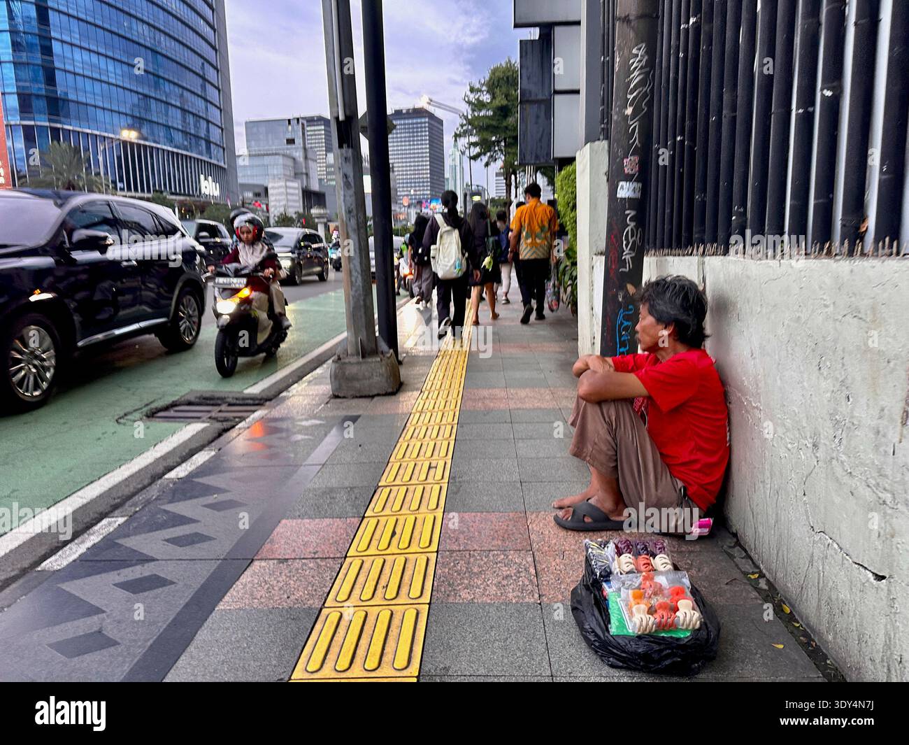 Jakarta - 5 mars 2026 : Un vendeur de rue est assis sur un trottoir vendant des collations emballées à côté de la circulation animée pendant que les piétons et les motos passent au crépuscule. - Image de stock capturée avec un smartphone