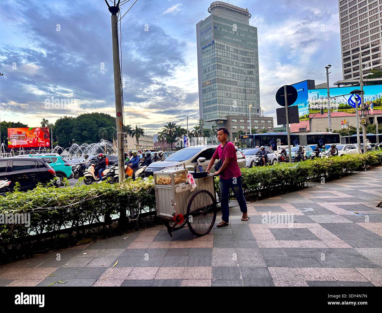 Jakarta - 5 mars 2026 : Un vendeur de rue pousse un chariot de nourriture le long d'un trottoir près de Bundaran HI à côté de la circulation très fréquentée. - Image de stock capturée avec un smartphone
