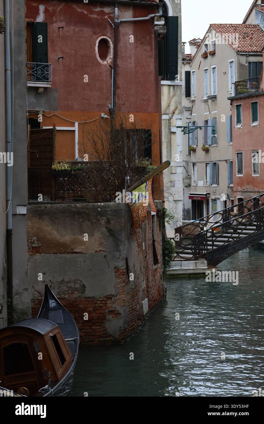 Coin de canal avec des bâtiments érodés, un bateau amarré et un pont décoratif dans un quartier calme de Venise. Banque D'Images