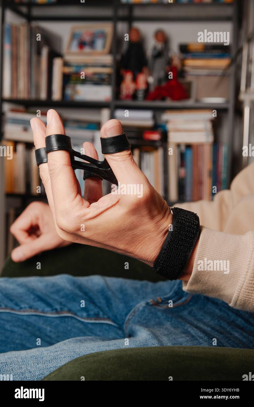 dans un salon, un homme exerce sa main avec un entraîneur de résistance des doigts, écartant ses doigts contre des bandes élastiques pour renforcer les muscles et le fle Banque D'Images