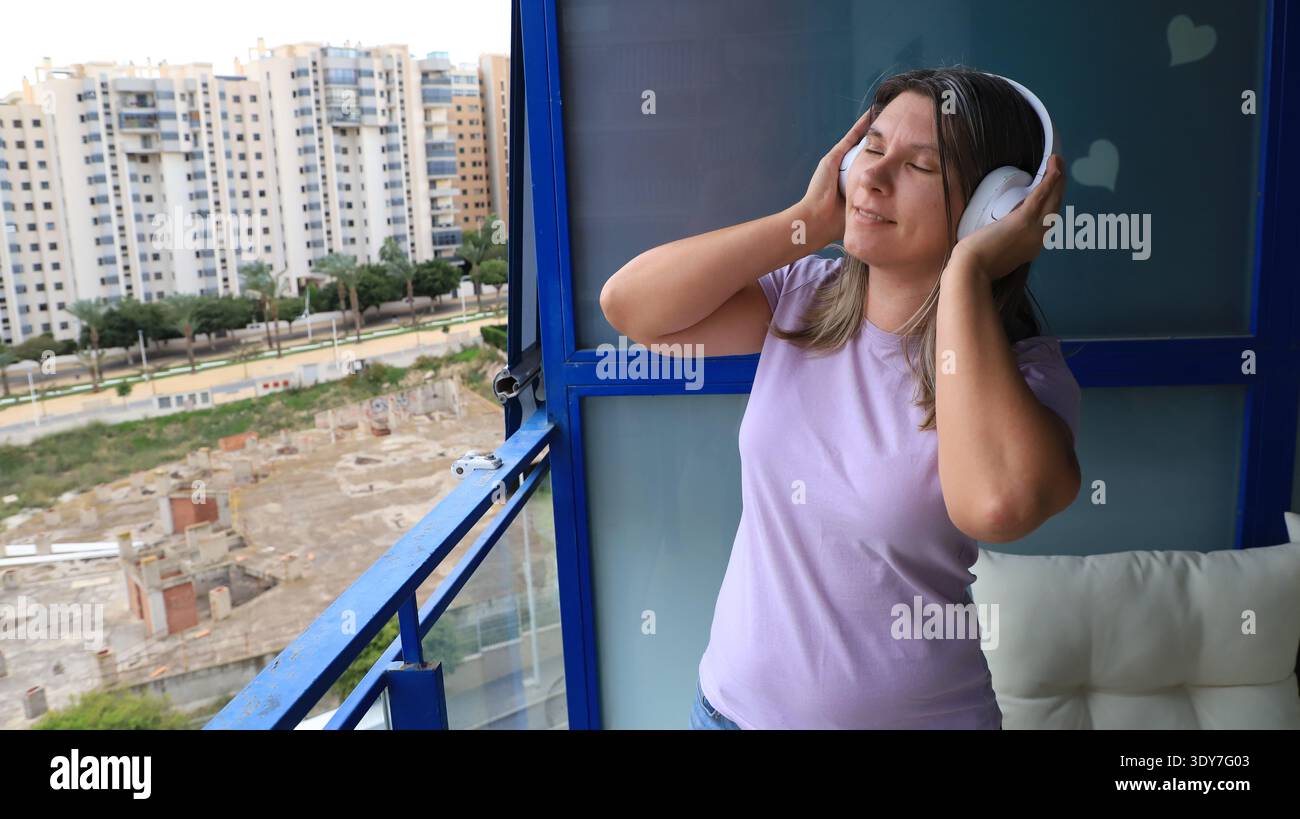 Femme portant des écouteurs aime la musique sur un balcon avec vue sur les bâtiments urbains Banque D'Images