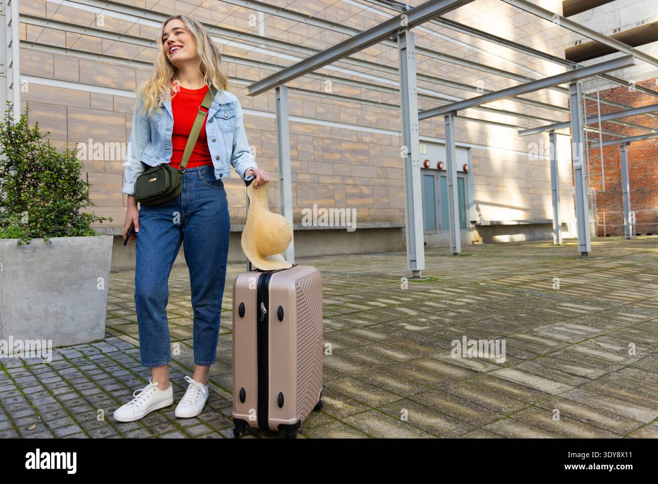Femme souriant dans la veste en denim à côté de la valise avec chapeau de paille, tenant le téléphone dans la plaza, espace de copie Banque D'Images