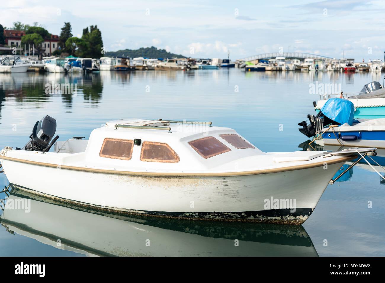 Petit bateau à moteur blanc vieilli avec moteur hors-bord flottant dans les eaux calmes du port avec une marina bondée. Banque D'Images