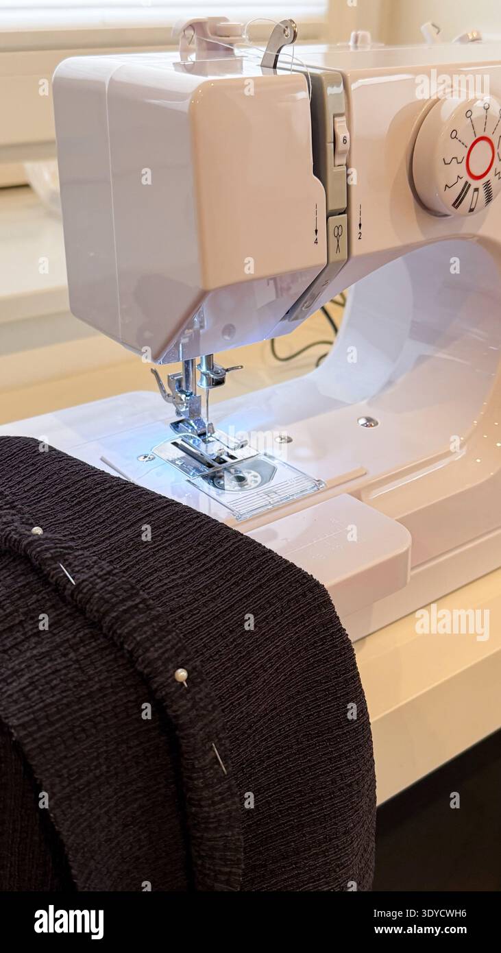 Machine à coudre avec tissu noir et épingles positionnées sur une table blanche, présentant l'aiguille et le pied-de-biche dans un espace de travail intérieur bien éclairé Banque D'Images