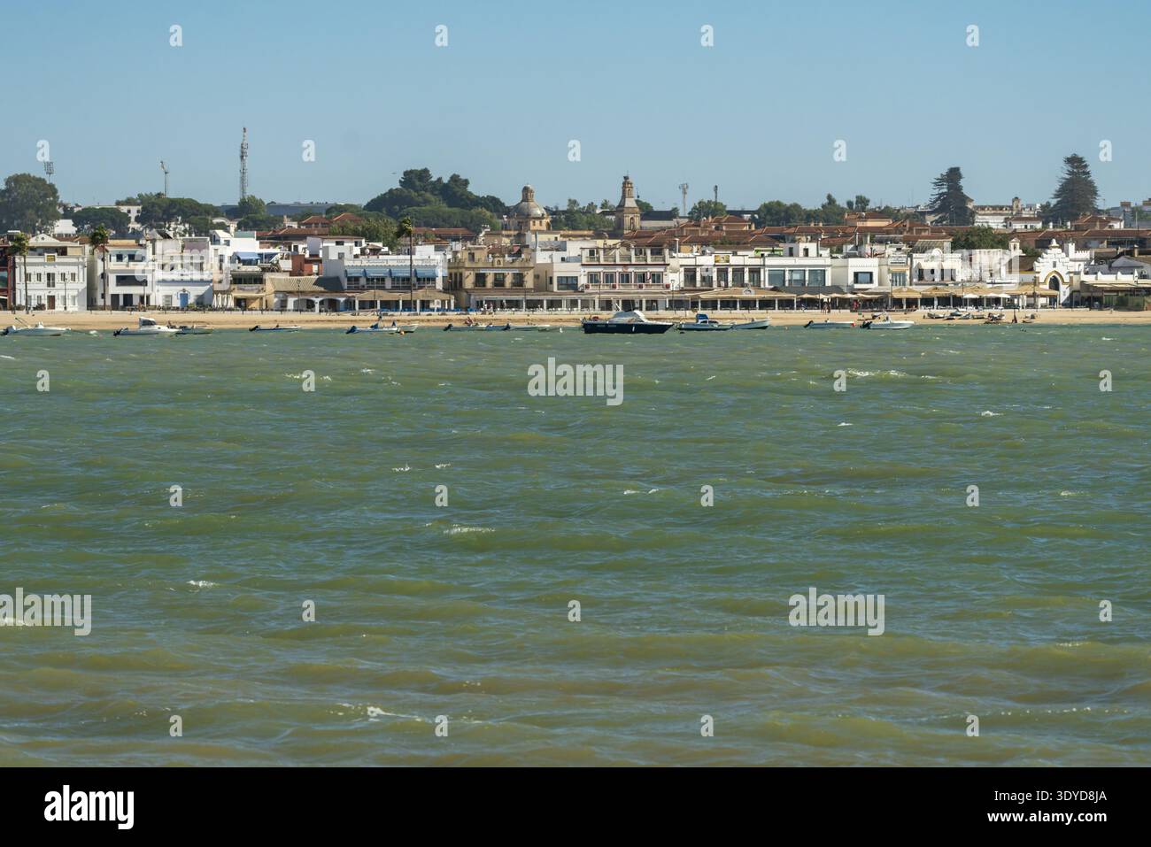 Scène de front de mer montrant une ville côtière de Sanlucar de Barrameda avec des bâtiments blancs, des églises et des petits bateaux sur la mer verdâtre sous ciel clair Banque D'Images