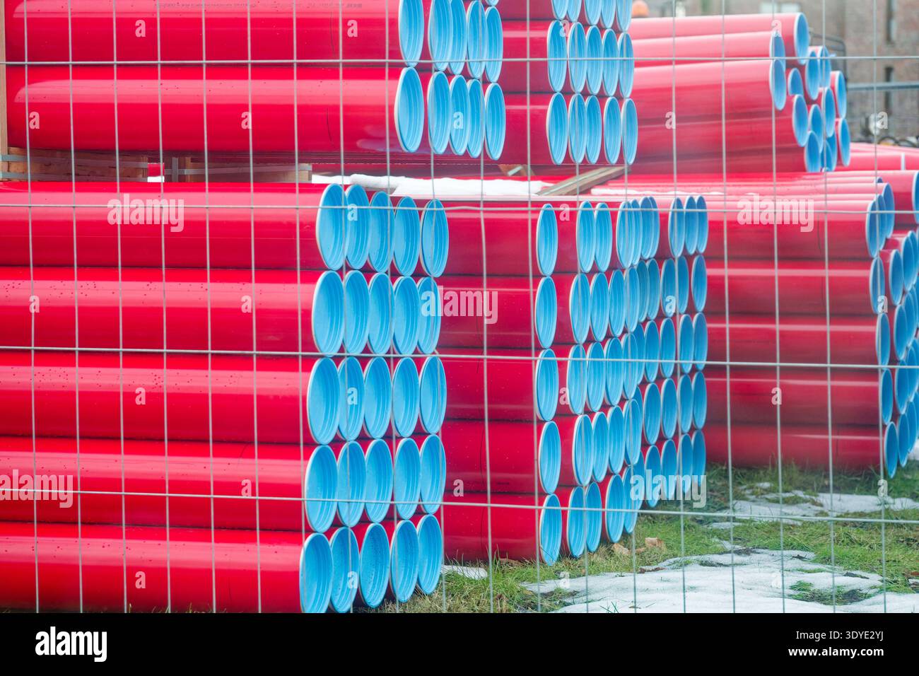 Les tuyaux en plastique rouge avec des extrémités bleues sont empilés en rangées organisées derrière une clôture métallique sur une zone extérieure herbeuse avec des taches de neige. Banque D'Images