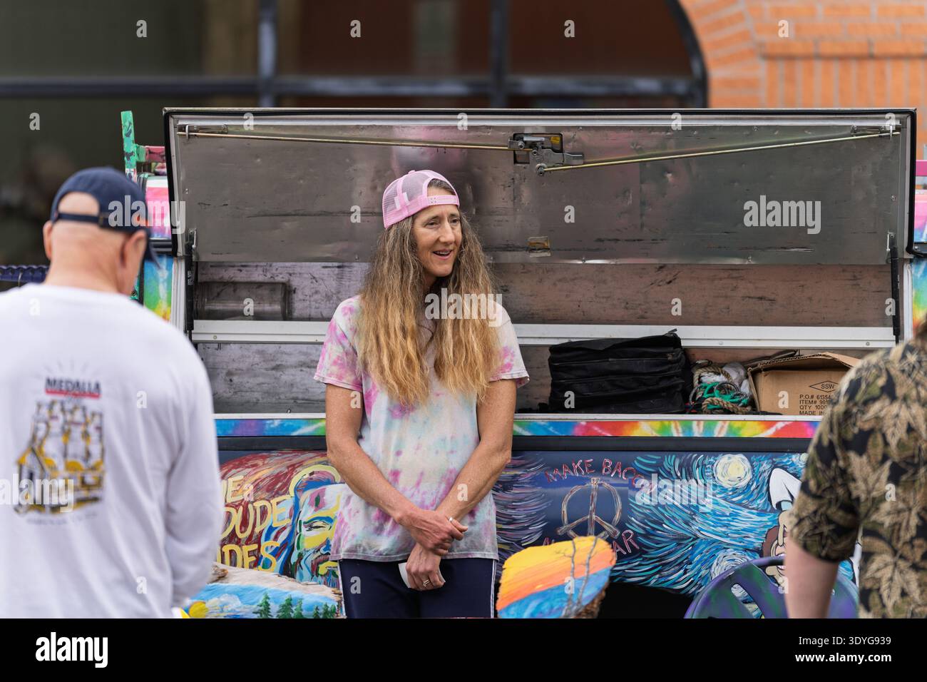 Une femme portant un chapeau de camionneur rose et une chemise teintée sourit tout en assistant à son vibrant camion de vendeur couvert d'art à Panama City, en Floride. Elle est PA Banque D'Images