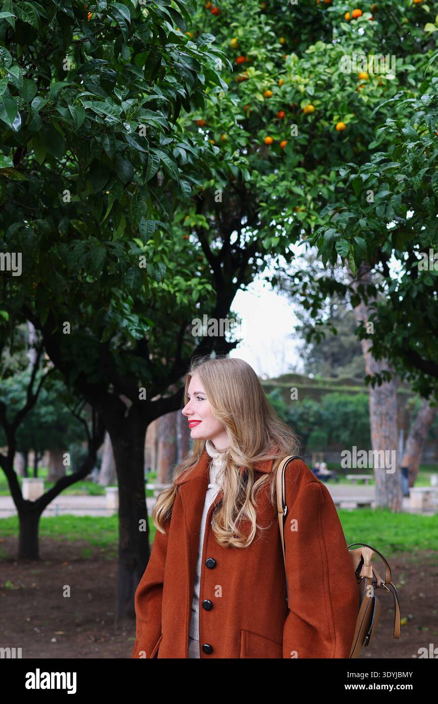 Femme élégante en manteau brun marchant dans un parc avec des orangers. Concept de style de vie et de voyage d'automne. Banque D'Images