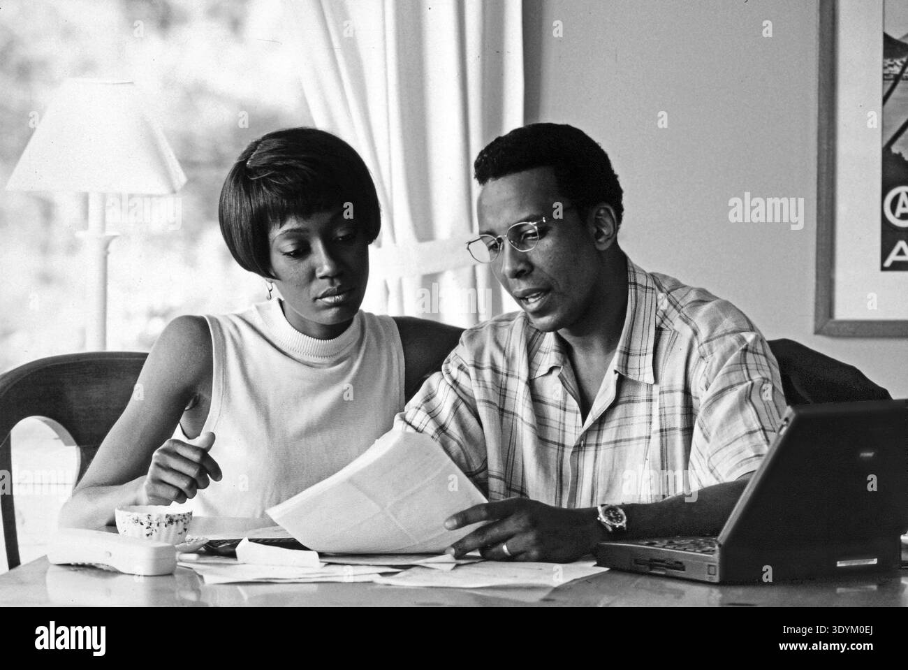Un couple afro-américain est assis à une table avec des papiers et des factures étalés. Ils se concentrent sur les documents tout en partageant leurs réflexions. Un ordinateur portable est ouvert à proximité Banque D'Images