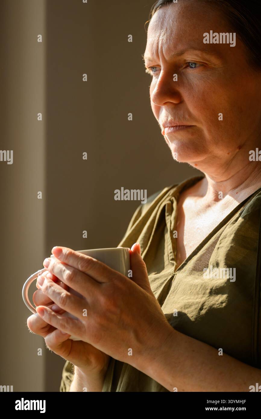 Femme d'âge moyen réfléchie tenant une tasse de thé par fenêtre dans la lumière chaude du matin, moment calme de réflexion à la maison. mise au point sélective Banque D'Images