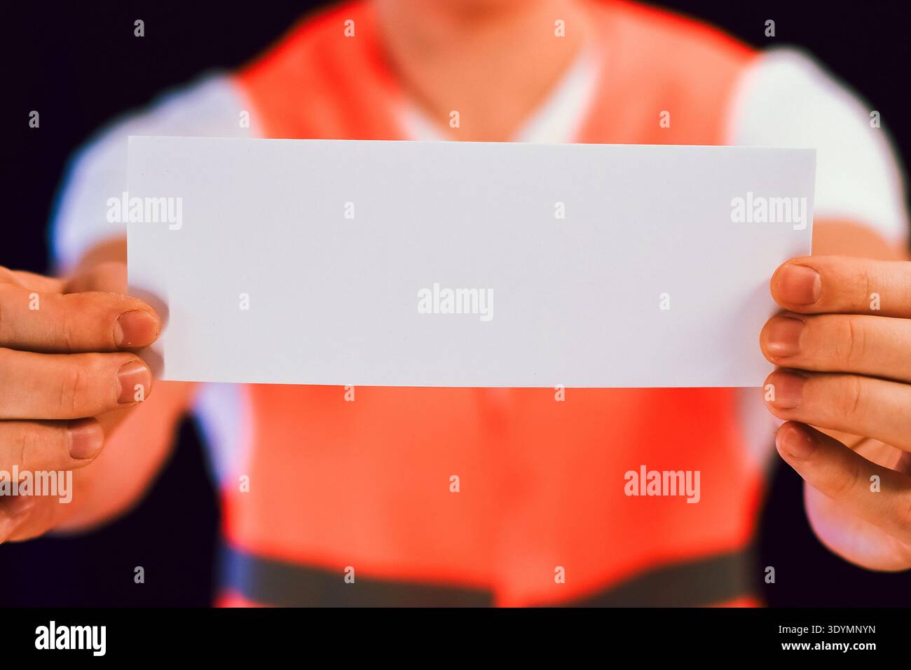 Vue rapprochée extrême des bras de la personne en gilet réfléchissant orange prolongeant la large feuille rectangulaire blanche vierge vers le spectateur sur noir Banque D'Images