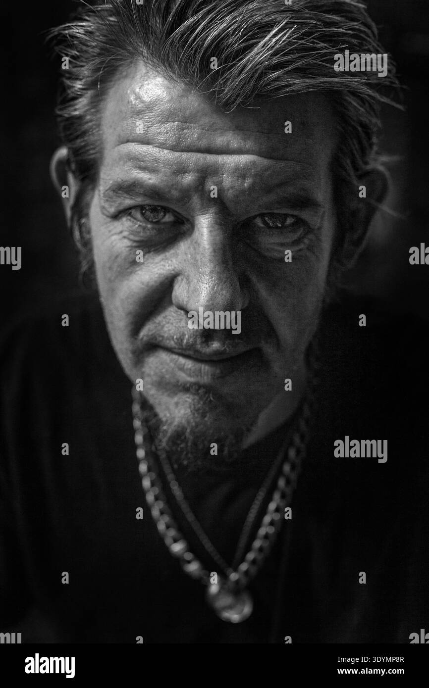 Gros plan gros plan portrait noir et blanc d'un homme mûr avec un bouc, des cheveux courts texturés et un regard intense, portant un collier en chaîne argentée. Banque D'Images