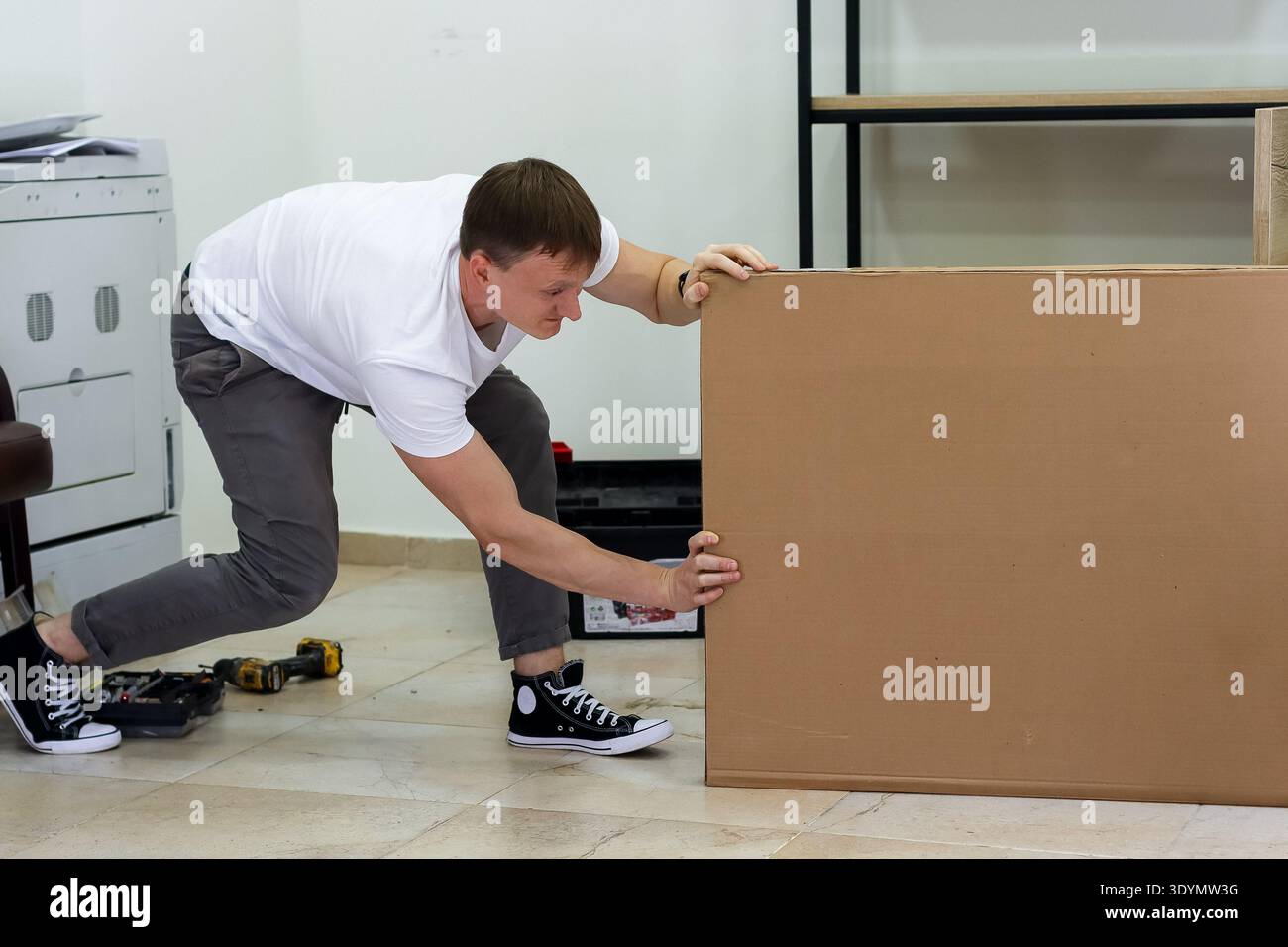 Homme poussant une lourde boîte en carton sur le sol. Homme déterminé en t-shirt blanc poussant une boîte en carton surdimensionnée tout en assemblant des meubles à la maison Banque D'Images