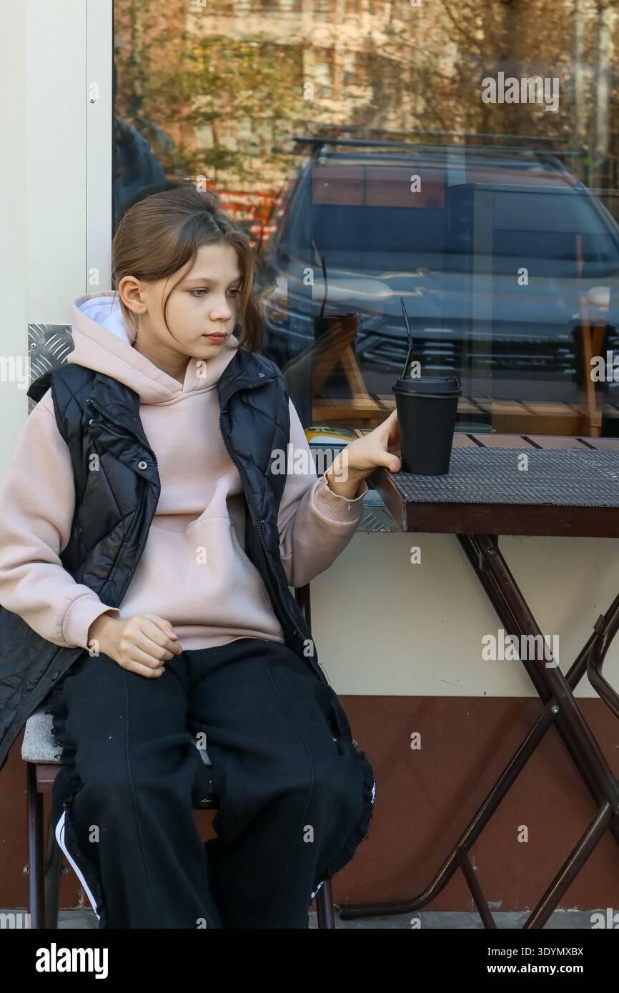 Preteen Girl en sweat à capuche beige et gilet noir assis au café de la rue, tenant une tasse de café noire à emporter, expression neutre, lumière naturelle Banque D'Images