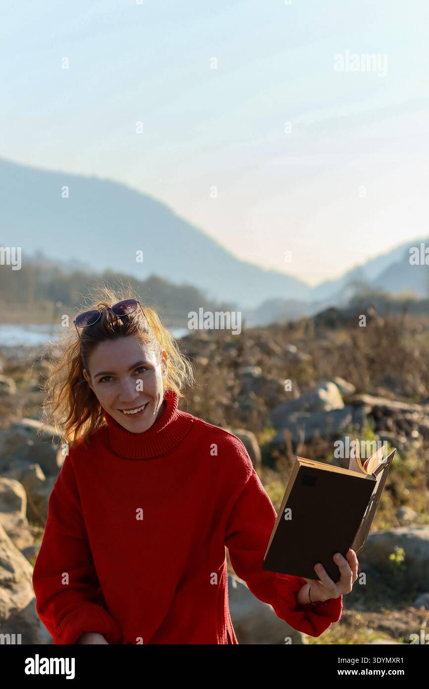 Heureuse jeune femme en pull rouge sourit tout en tenant un livre ouvert dans un cadre extérieur rocheux avec des montagnes brumeuses en arrière-plan. Banque D'Images