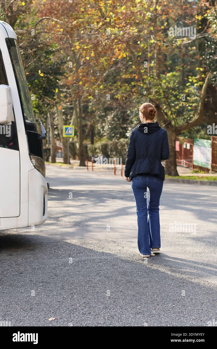 Plan large d'une jeune femme en tenue décontractée quittant un arrêt de bus blanc sur une route bordée d'arbres d'automne vibrants et d'herbe. Banque D'Images