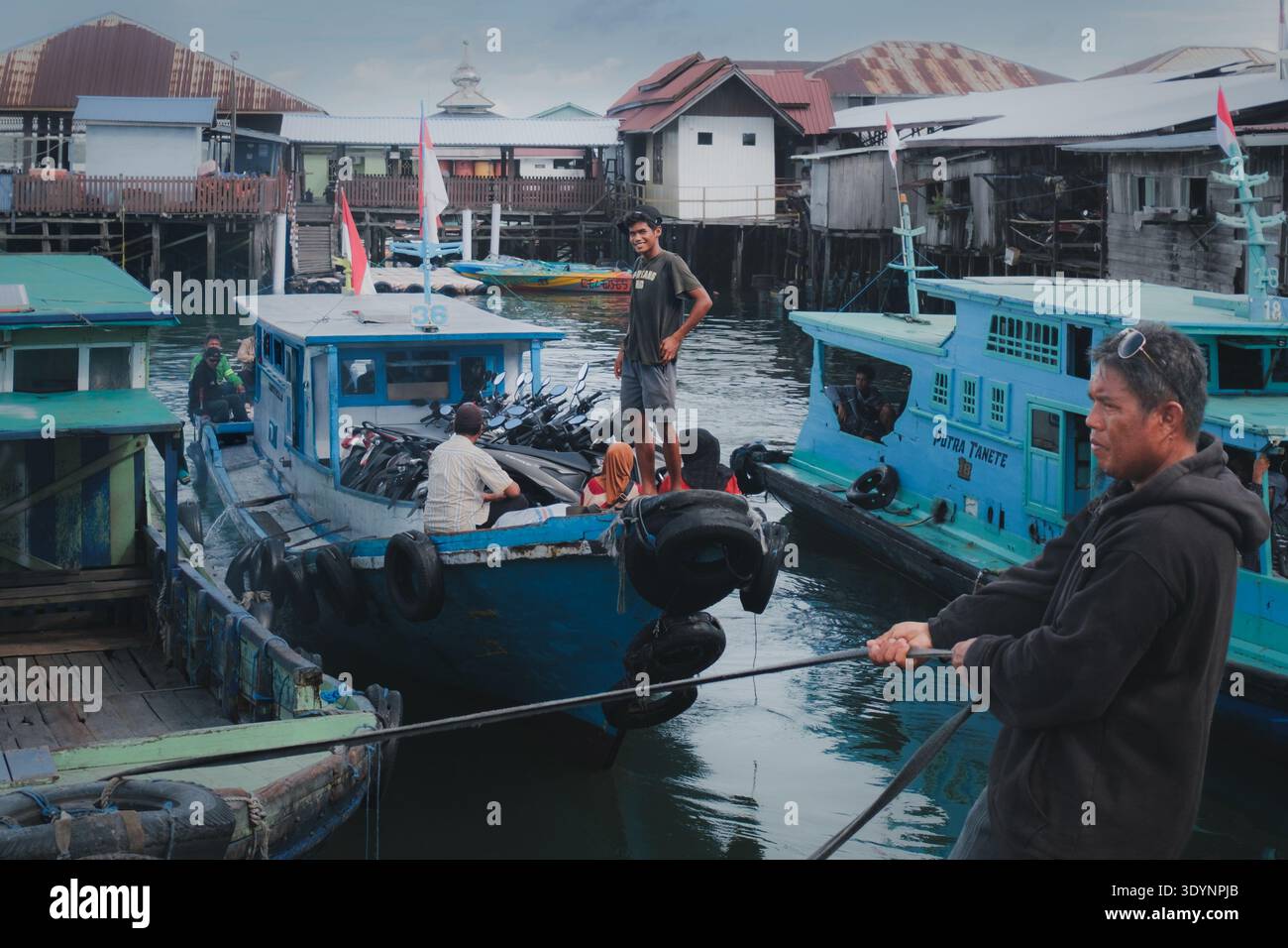 Balikpapan, Indonésie - 21 décembre 2025. Un ferry klotok en bois bleu transporte des passagers et une rangée de motos à travers l'eau dans un terminal très fréquenté. Banque D'Images