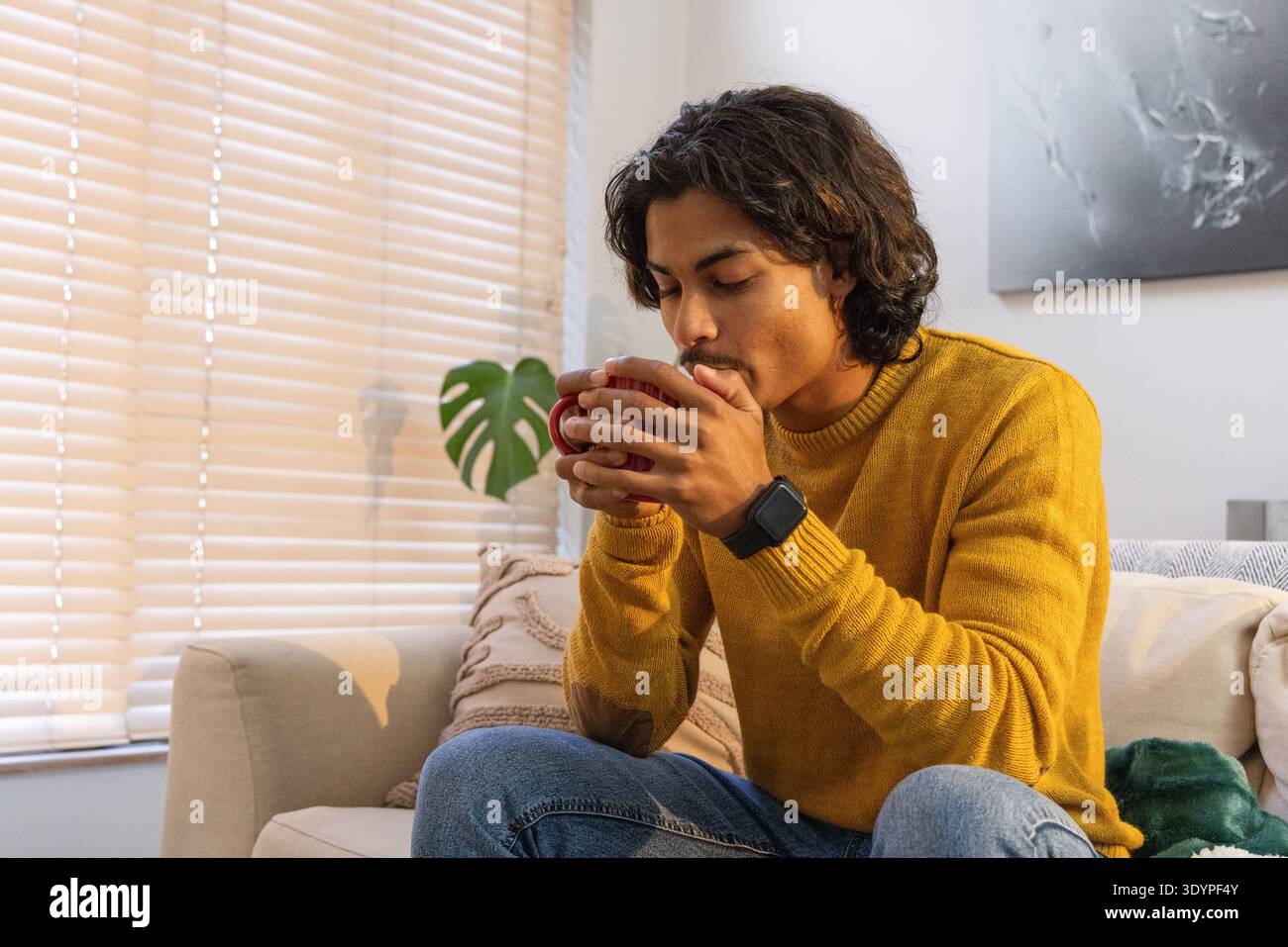 Homme asiatique portant un pull moutarde assis sur un canapé dans le salon tenant la tasse rouge smartwatch visible Banque D'Images