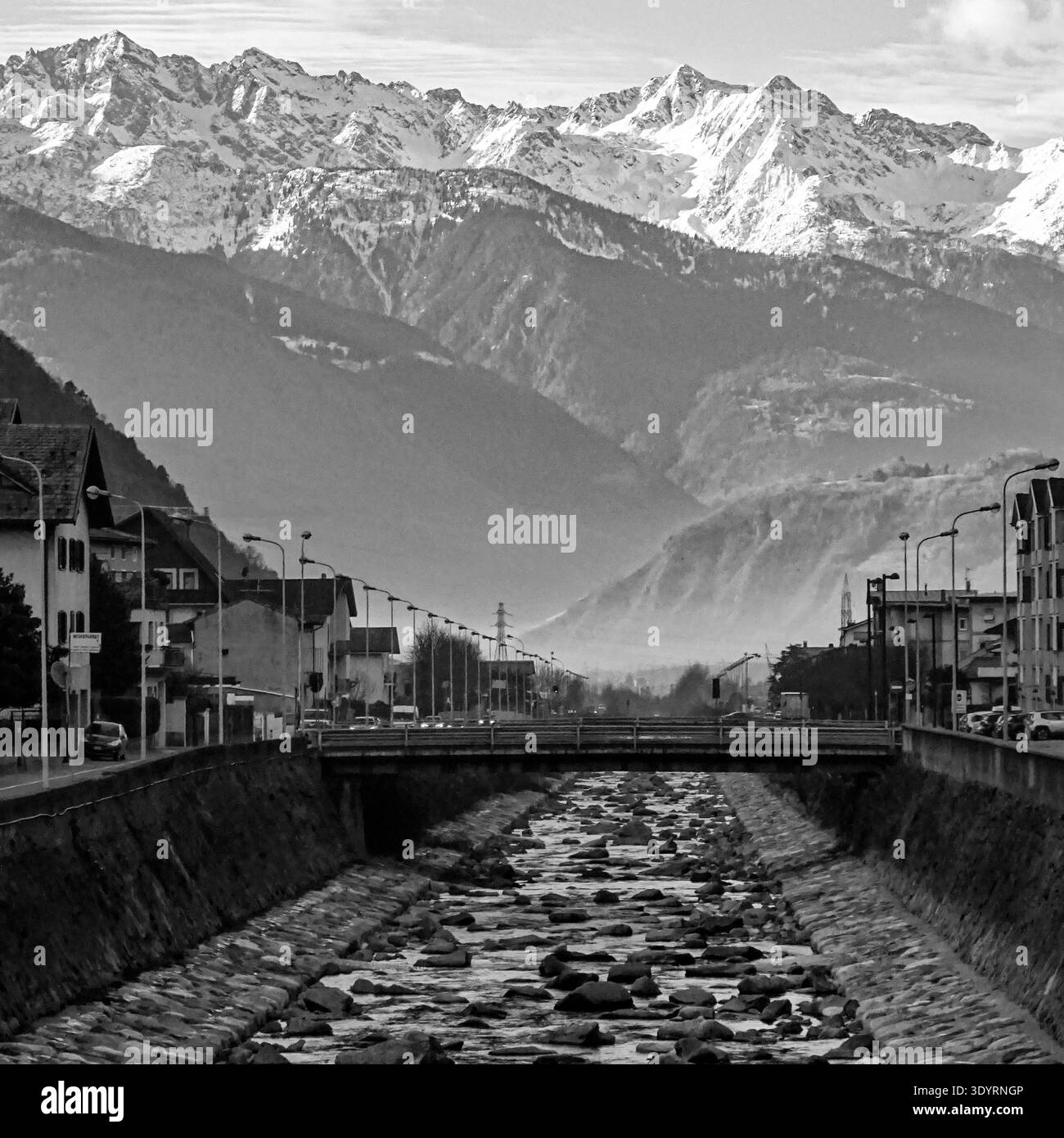 Vue en noir et blanc d'une ville avec canal et pont, avec des montagnes enneigées en arrière-plan. Banque D'Images
